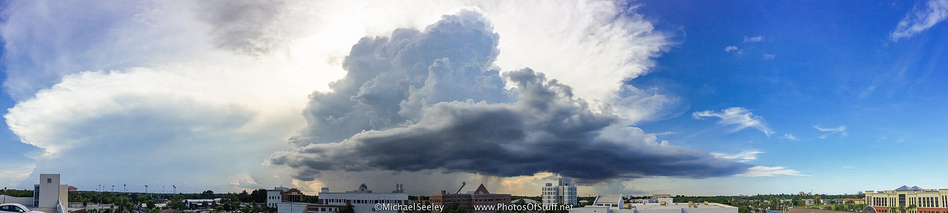 Storm clouds over the Florida Institute of Technology campus in Melbourne, with academic buildings, palm trees, and a green lawn visible beneath dramatic skies.